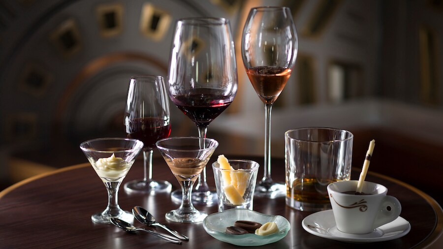 7 glasses containing various types of liquor next to a glass and a dish containing chocolate