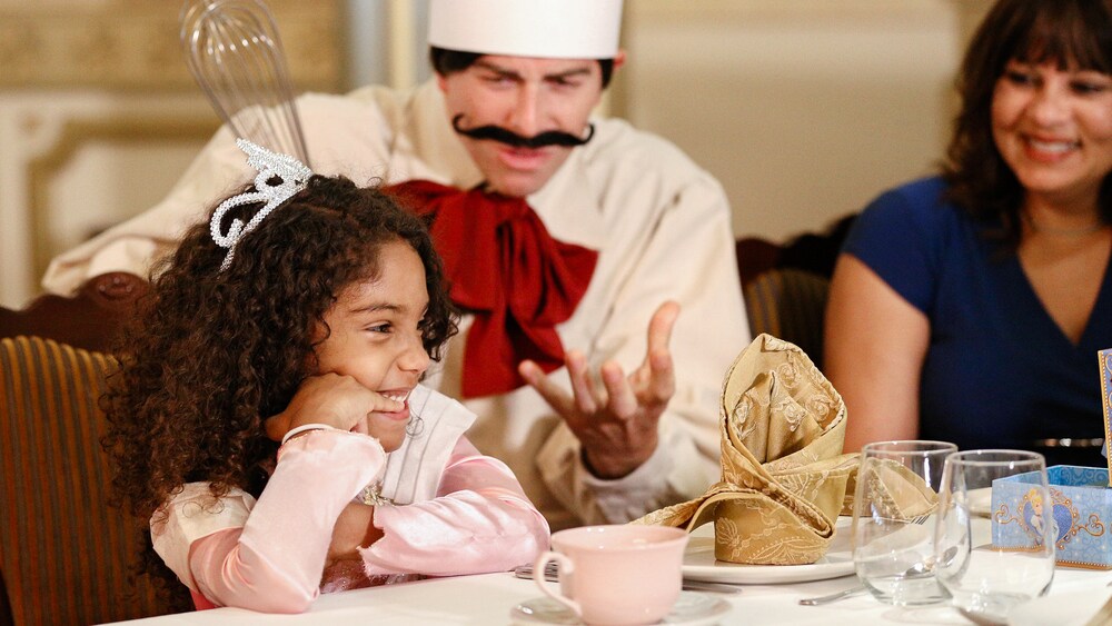 A little girl, wearing a tiara, sits next to her mother and laughs while a Royal Pastry Chef tells stories