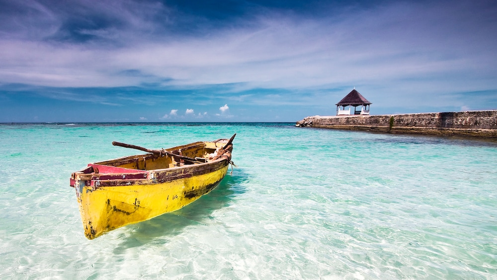 An old rowboat floats in clear waters next to a breaker with a small gazebo structure