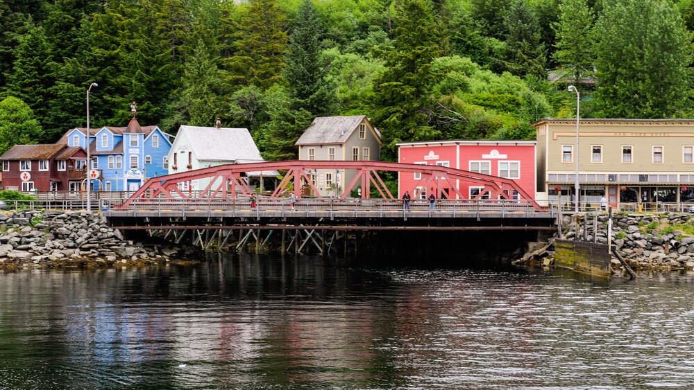 A bridge extends over a small inlet with buildings and woods in the background