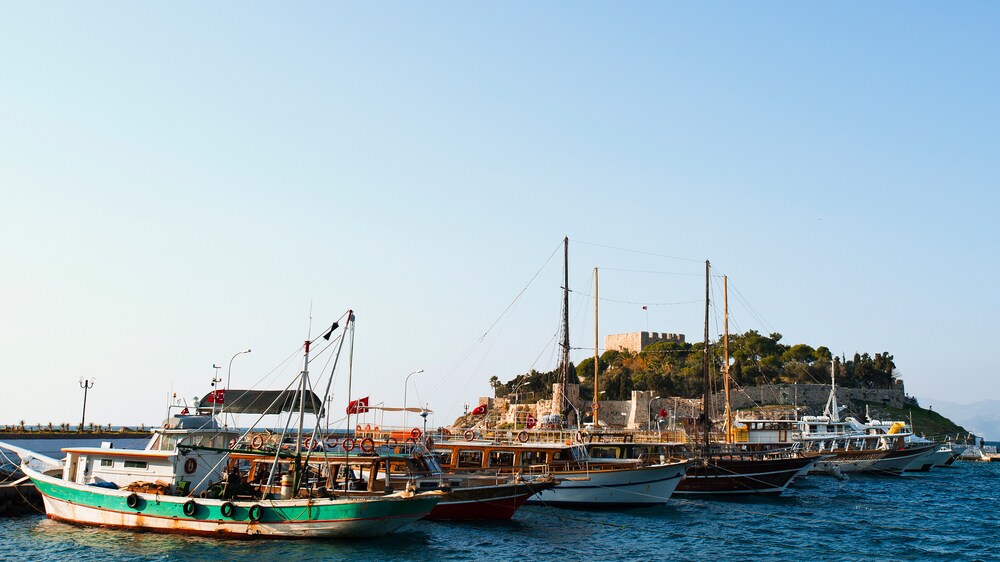 Six fishing boats bearing the Turkish flag sit in a row in front of a round island circled by stone walls