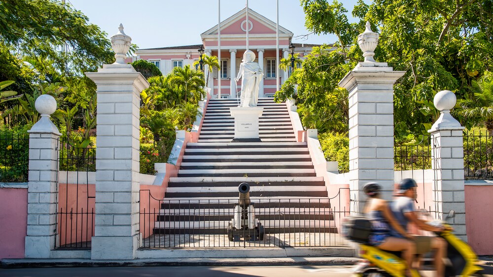 Behind a low fence and a cannon, stairs lead up past a statue to a large house with columns