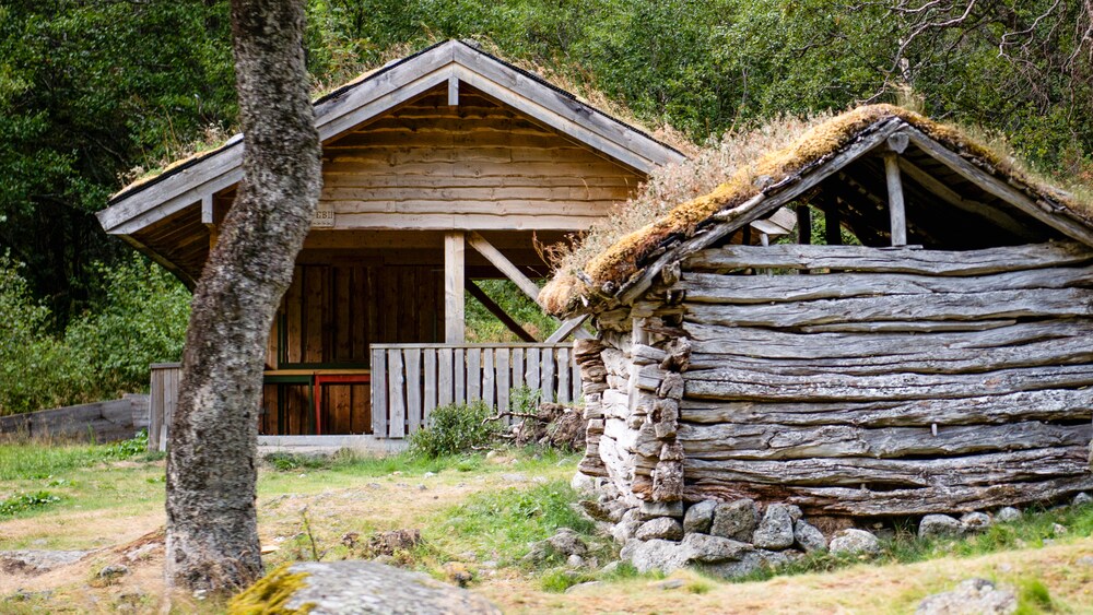 A building with a raised roof sits behind a smaller structure built of interlocking logs