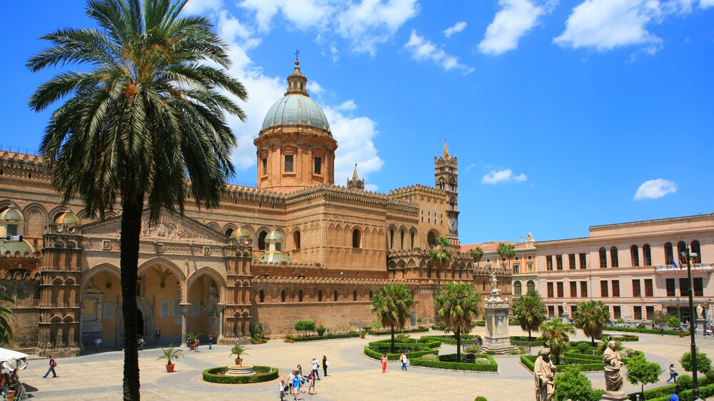 People walking along a terrace before a large building with windows and a rooftop cross
