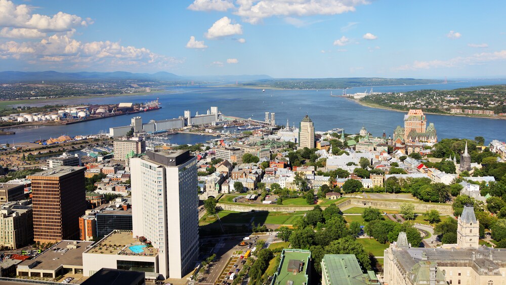 A city filled with trees, buildings and a crowded harbor near a populated body of water set against a backdrop of verdant hills