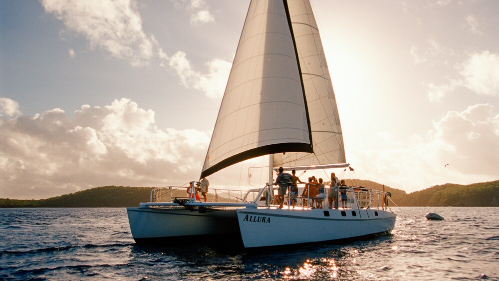 Several people relax on a catamaran with its sails up as it floats on the ocean at twlight