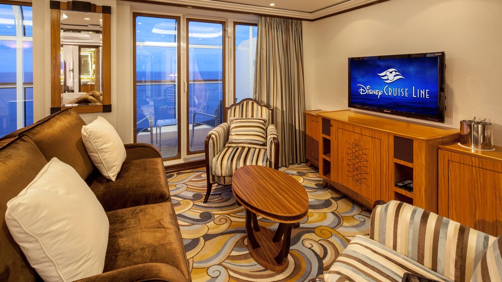 A Disney Cruise Line Concierge 1 Bedroom Suite with Verandah Stateroom with a sofa, a verandah with an ocean view, 2 chairs, a low table, a cabinet and a TV