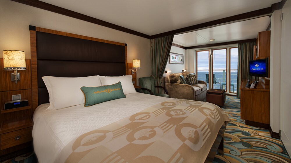 A Disney Cruise Line Concierge Verandah Stateroom with a bed, a sleeper sofa, a low table, a verandah with an ocean view, a desk, shelves and a TV