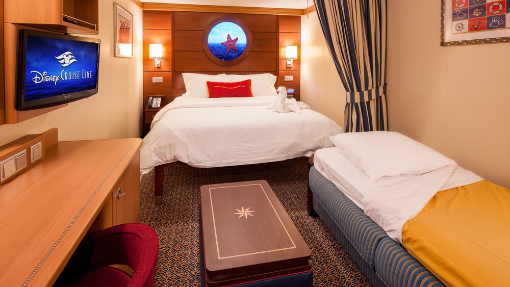 A Disney Cruise Line Standard Inside Stateroom with a desk, a TV, a bed, a virtual porthole, a sleeper sofa and a low table