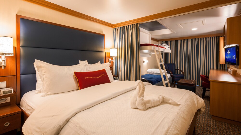 A Disney Cruise Line Deluxe Family Oceanview Stateroom with a bed, a bunk bed, a chair, a TV, storage shelves and a desk 