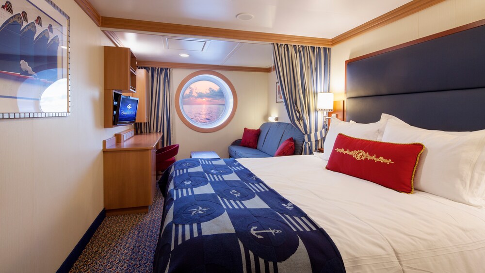 A Disney Cruise Line Deluxe Oceanview Stateroom with a desk, storage shelves, a porthole with an ocean view, a sofa and a bed