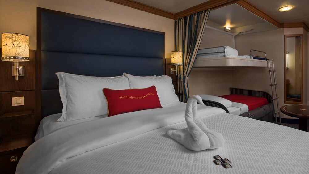 A Disney Cruise Line Deluxe Inside Stateroom with a bed, mirror, table, 2 bunk beds and a towel folded into the shape of a swan