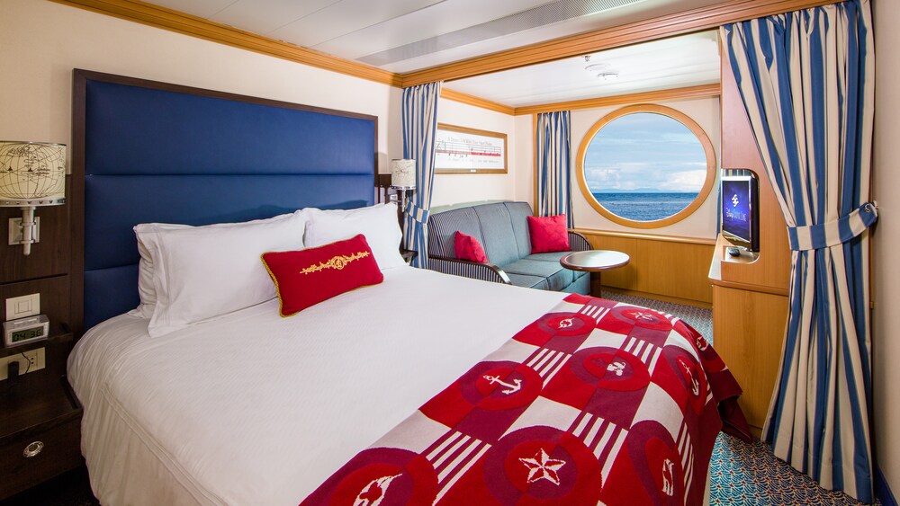 A Disney Cruise Line Deluxe Oceanview Stateroom with a bed, sofa, table, desk, nightstand, TV and a window facing the ocean