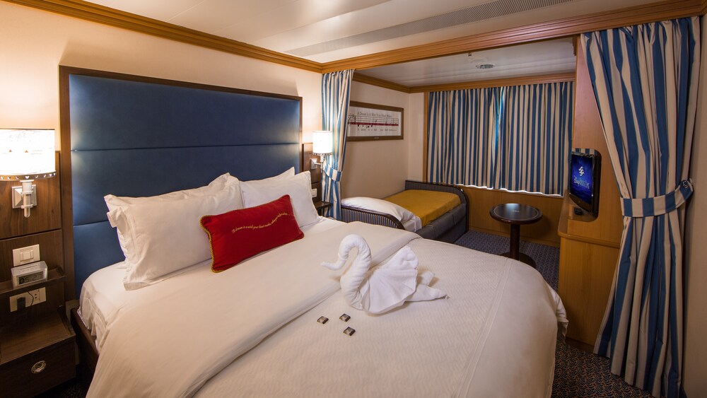 A Disney Cruise Line Deluxe Oceanview Stateroom with a bed, sofa bed, table, desk, TV, nightstand and a towel folded into the shape of a swan