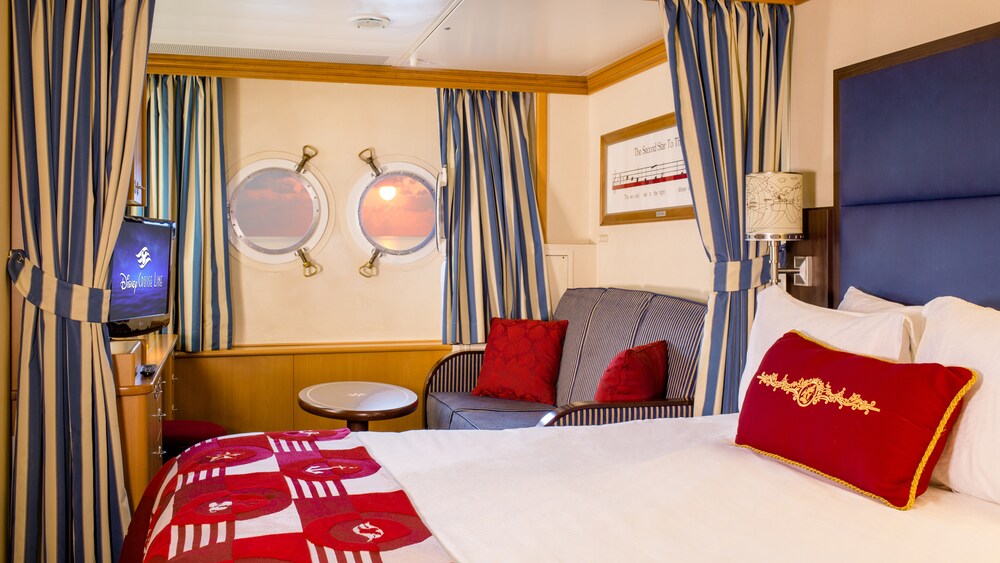 A Disney Cruise Line Deluxe Oceanview Stateroom with a bed, sofa, table, desk, TV and 2 windows facing the ocean