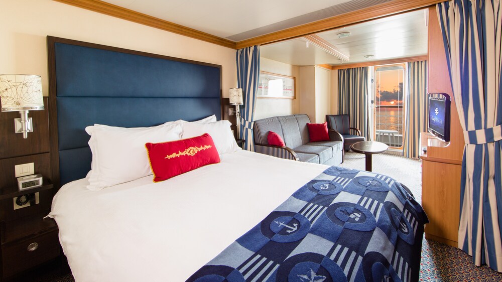 A Disney Cruise Line Deluxe Family Oceanview with Verandah Stateroom with a bed, sofa, chair, table, desk, TV, 2 nightstands and a verandah facing the ocean