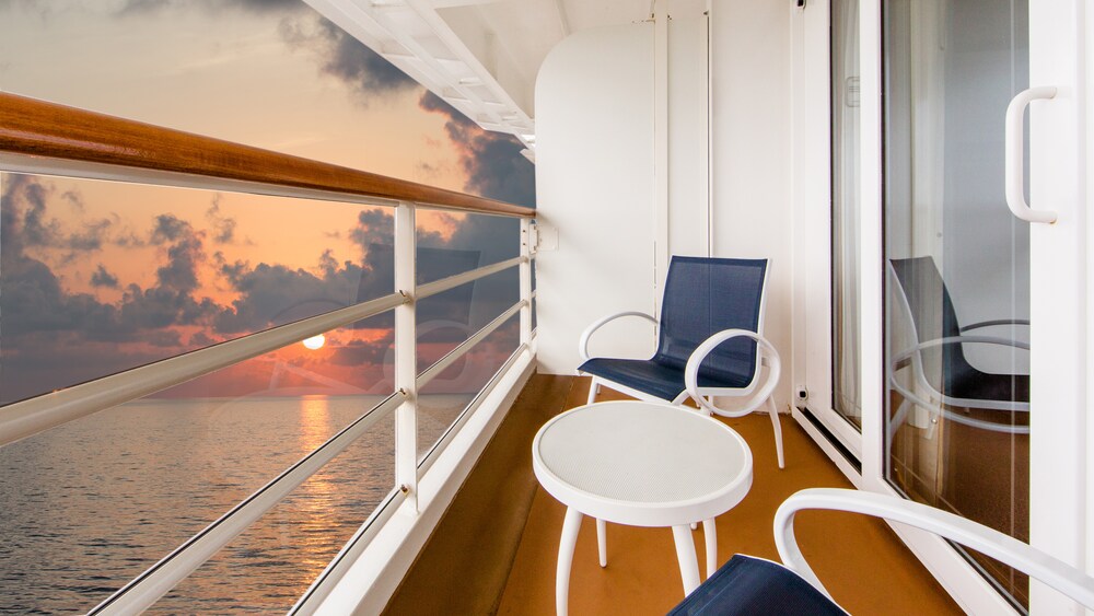 Two chairs and a table on a verandah, facing a plexiglass protected railing with an ocean sunset visible