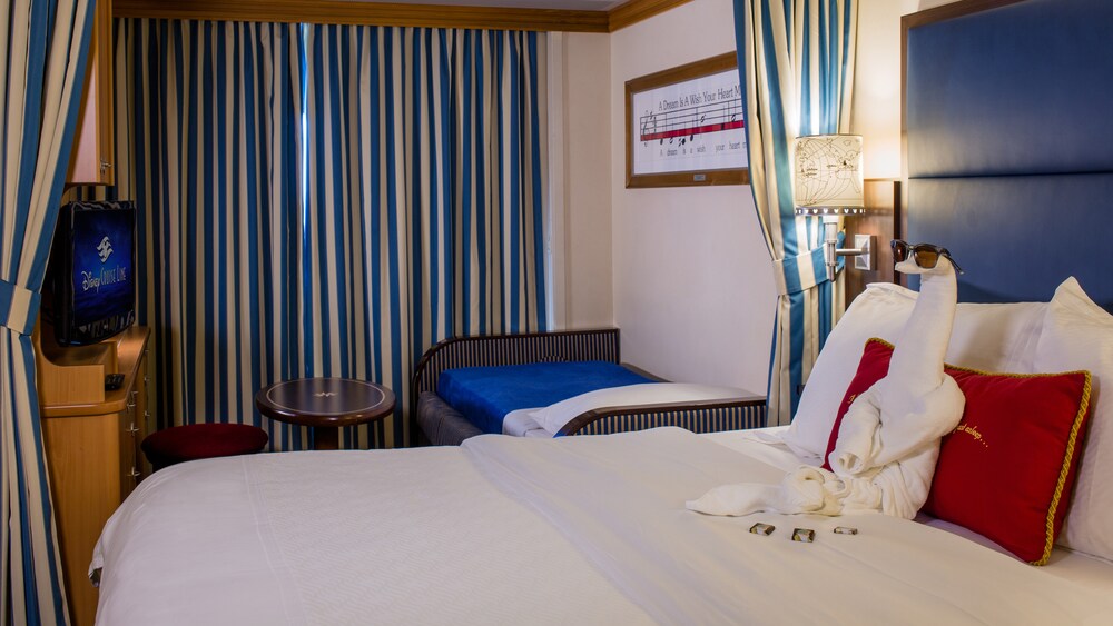 A Disney Cruise Line Oceanview with Verandah Stateroom with a bed, sofa bed, table, desk, TV and a towel folded into the shape of a swan
