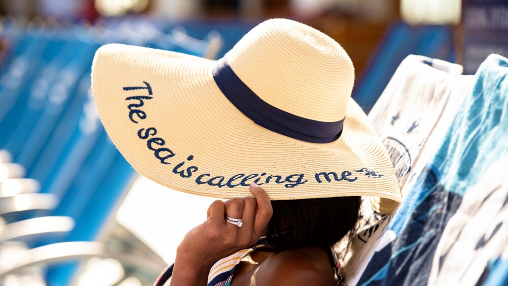 A woman sitting on a lounge chair and wearing a sun hat that reads ‘The sea is calling me’