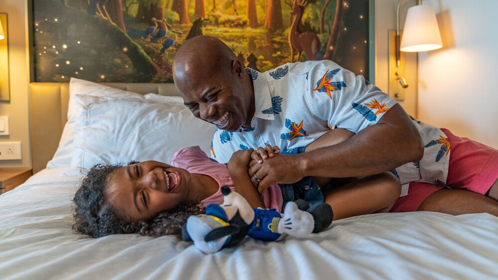 A father tickles his daughter on a bed while she laughs next to a Disney Cruise Line Mickey Mouse plush doll