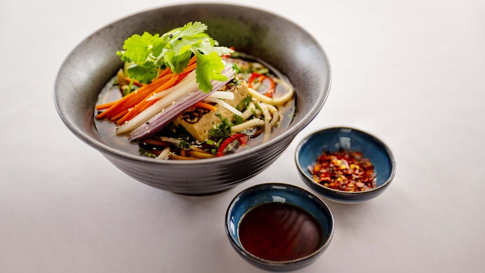 A bowl of noodle soup with tofu and fresh vegetables, with 2 cups of sauce and red chili flakes
