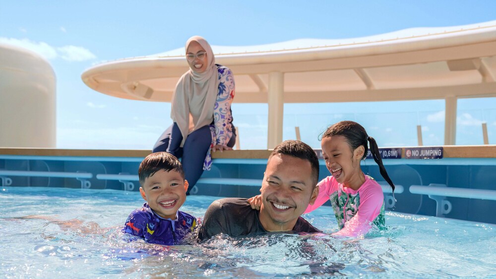 A father and his 2 young children playing in a pool while their mother sits on the edge of the pool
