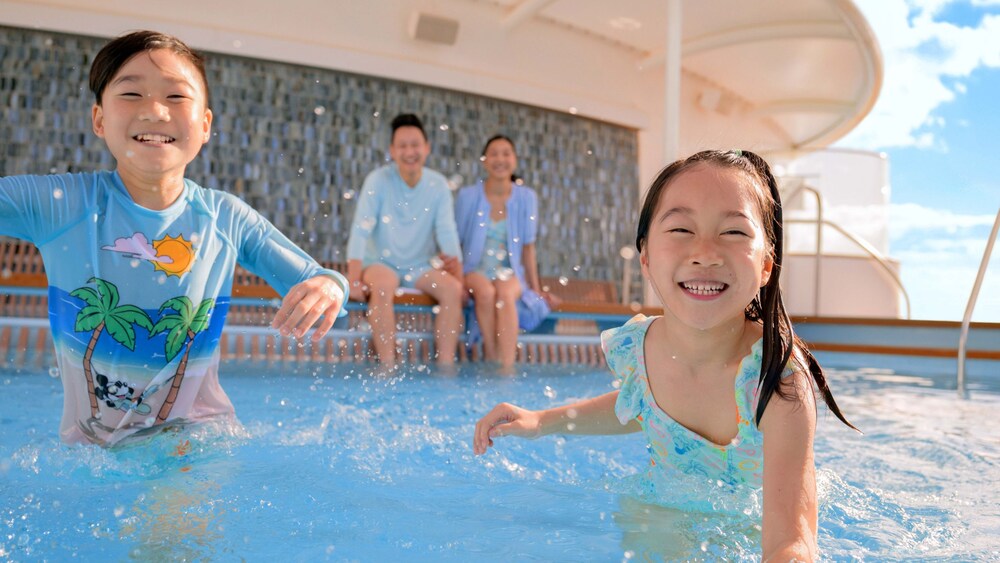 Two young children play in a pool while their parents sit on the pool's edge and watch