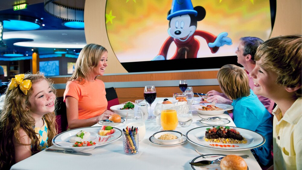 A family of 5 eating a meal at a dining table next to a screen with Mickey Mouse from 'Fantasia'