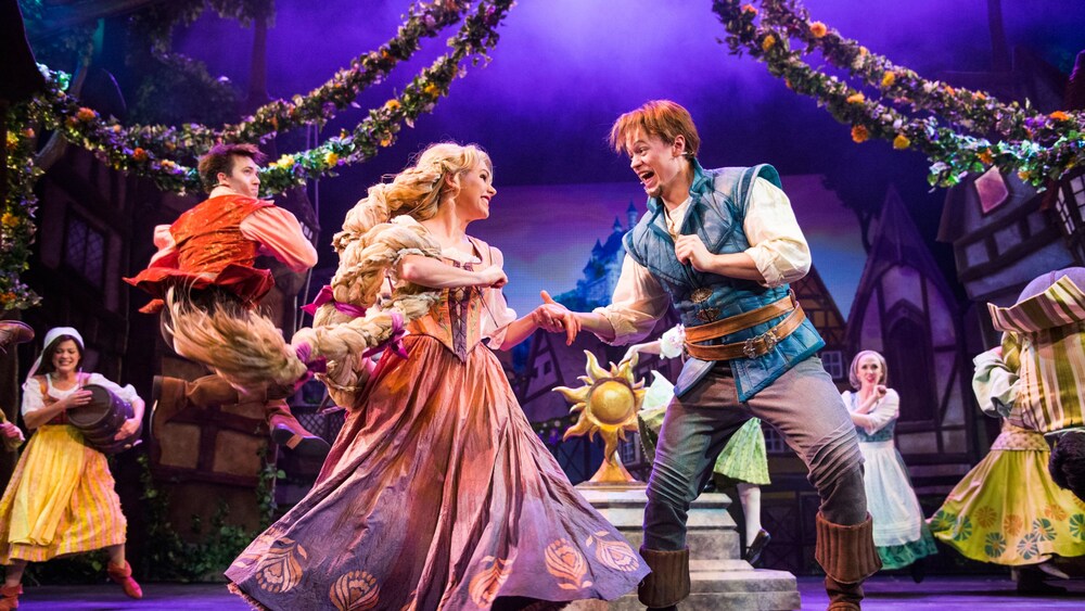 Rapunzel and Flynn Rider hold hands while dancing on stage in Tangled The Musical