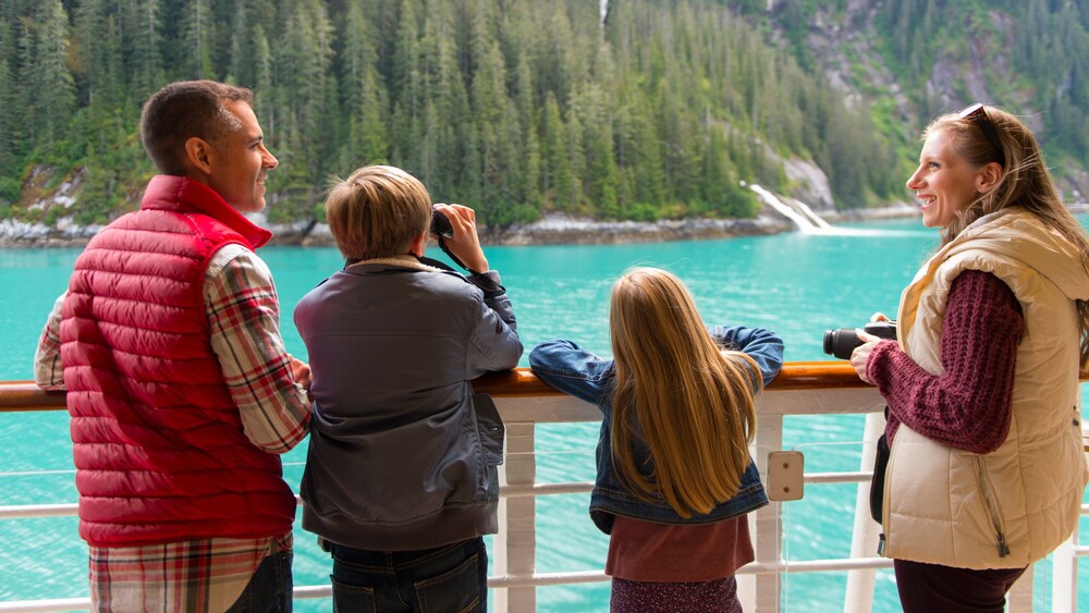 A family of 4 standing at the railing of a Disney Cruise Line ship and admiring a scenic fjord in Alaska