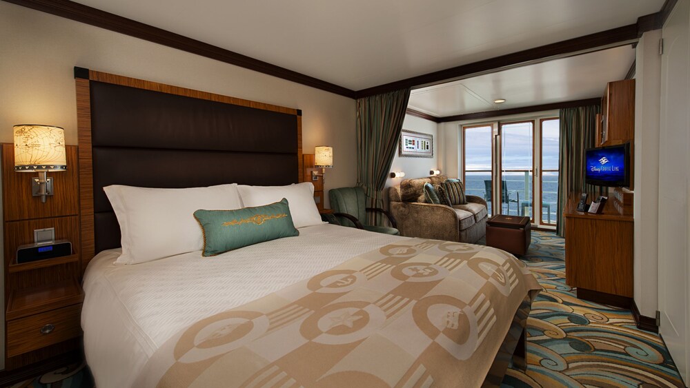 A Disney Cruise Line Concierge Verandah Stateroom with a bed, a sleeper sofa, a low table, a verandah with an ocean view, a desk, shelves and a TV