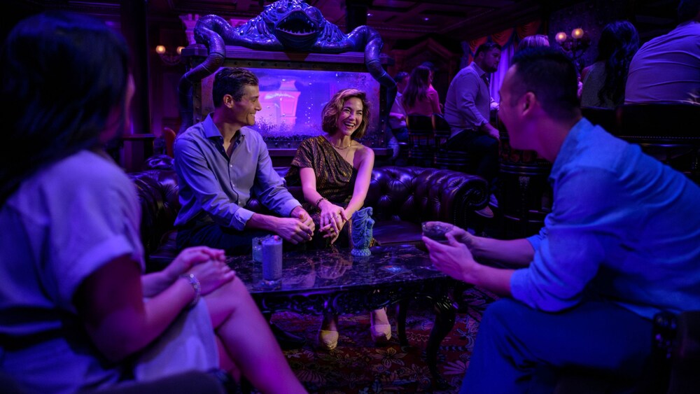 Cuatro Huéspedes sonríen y disfrutan de bebidas mientras están sentados alrededor de una mesa de Haunted Mansion Parlor en el Disney Treasure
