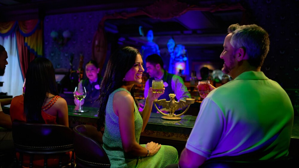 Cuatro Huéspedes disfrutando de bebidas mientras dos Miembros del Elenco preparan bebidas detrás del bar de Haunted Mansion Parlor en el Disney Treasure