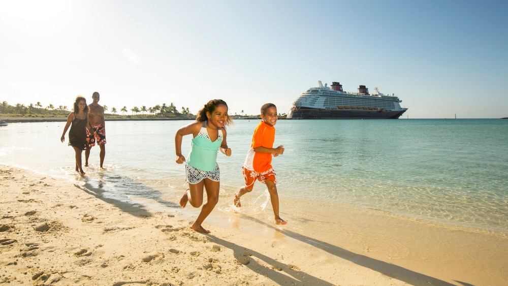 Two children running on the beach at Disney Castaway Cay with their parents walking behind and a Disney Cruise Line ship docked in the background