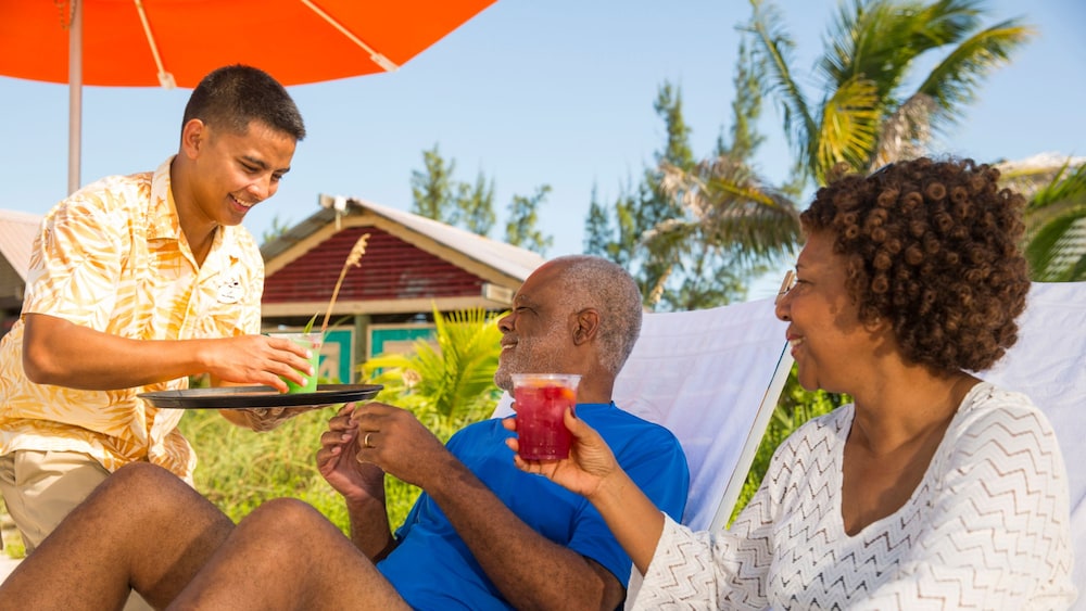 A Cast Member serving drinks to a couple relaxing under an umbrella at Serenity Bay on Disney Castaway Cay