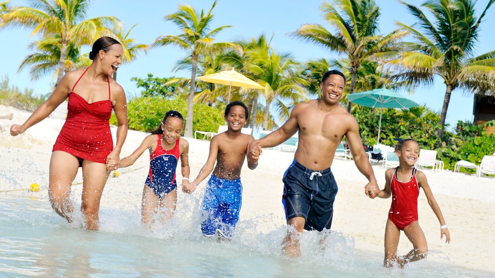 A family of 5 splashing in the shallow water at Disney Castaway Cay, with palm trees and beach umbrellas in the background