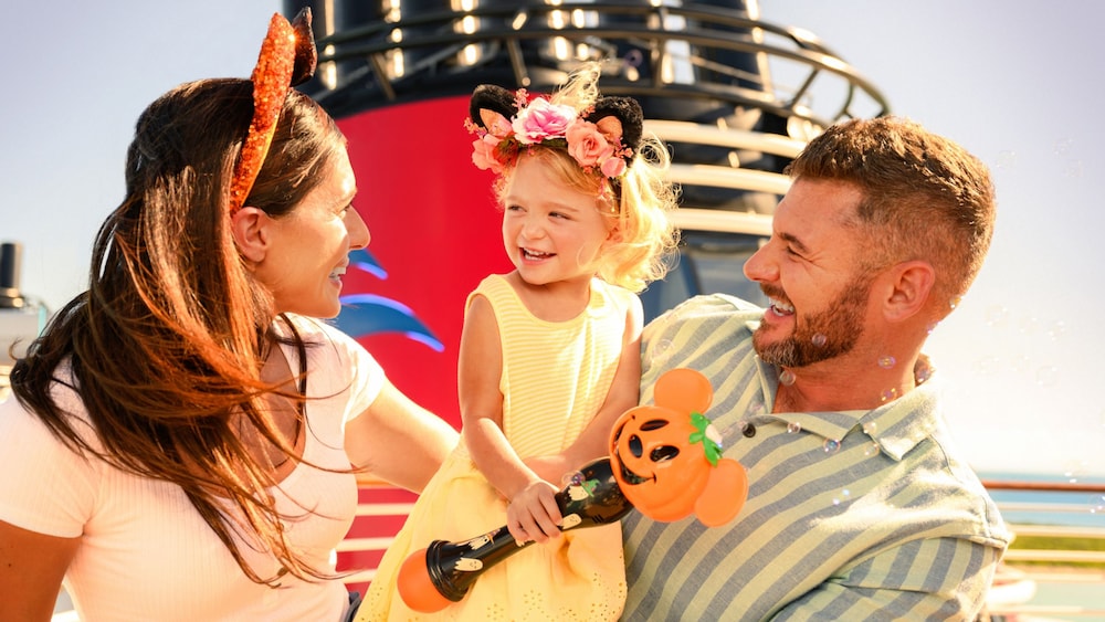 A couple carrying a young girl holding a bubble maker near a Disney Cruise Line ship funnel