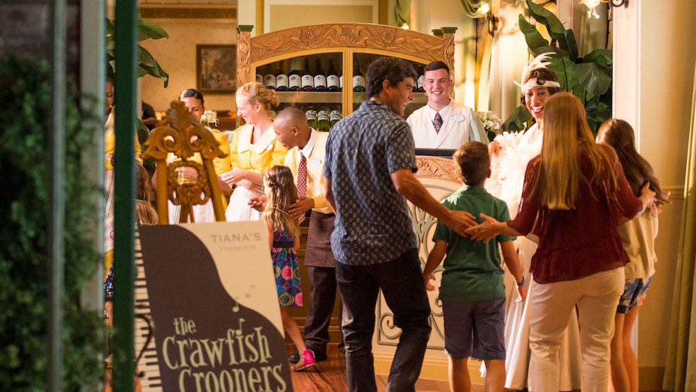 A family of 4 being greeted by Cast Members as they enter Tiana's Place, with a sign out front that says, 'The Crawfish Crooners'