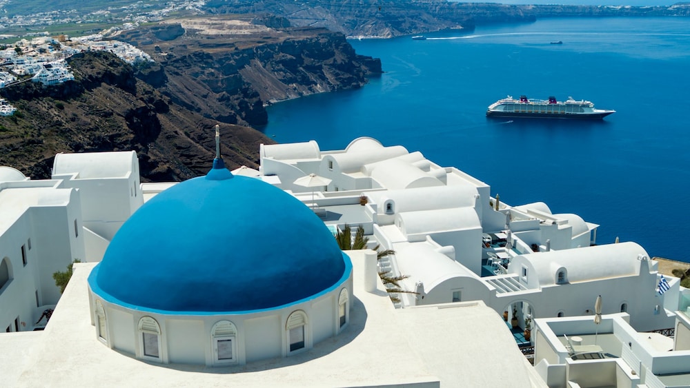 A blue domed church in Oia, Santorini, a cliffside and the sea with a cruise ship