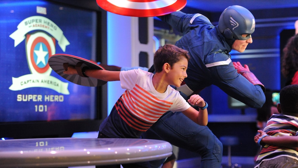 A child holding a shield and posing in a battle stance next to Captain America at Marvel Super Hero Academy