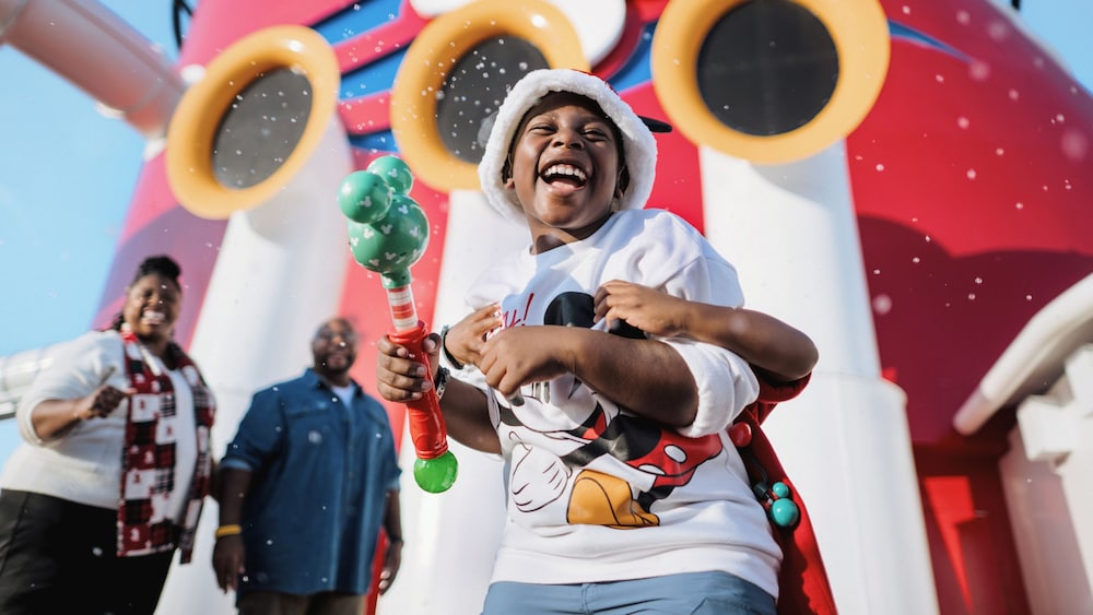 A child wearing a Santa hat laughs while being hugged from behind on the deck of a Disney Cruise Line ship