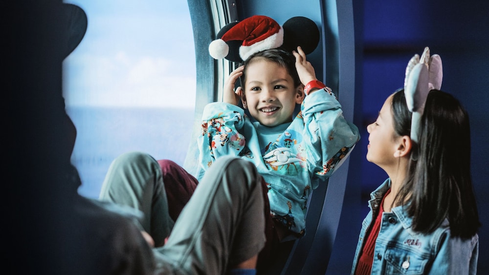 Uma garota sorri para o irmão enquanto ele se reclina em um assento próximo à escotilha e ajusta sua tiara de orelhas de Mickey Noel