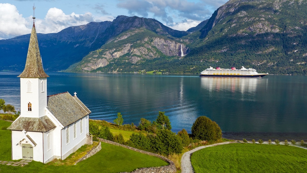 Uma antiga capela na margem de uma enseada, com um navio da Disney Cruise Line e montanhas ao fundo