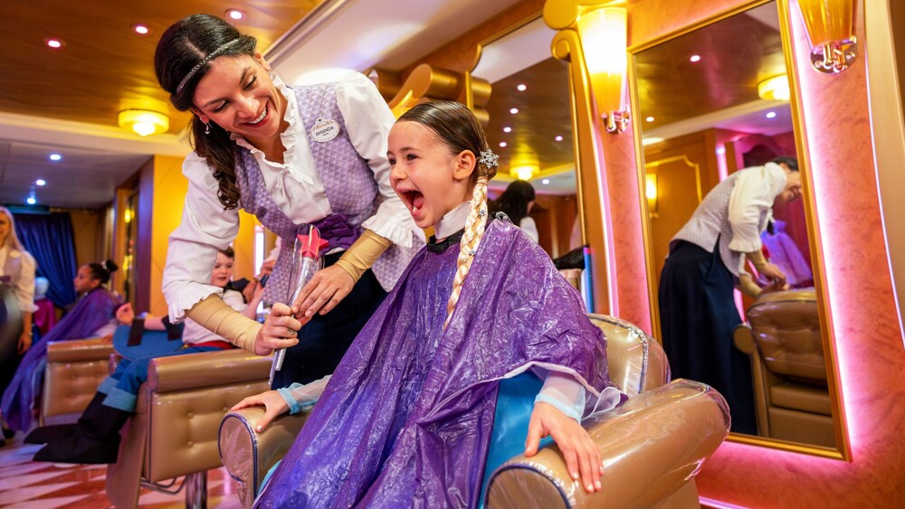 A young girl smiling in a salon chair while being served by a Cast Member at the Bibbidi Bobbidi Boutique