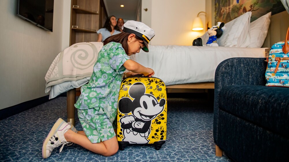 A young child kneeling and unzipping a Mickey Mouse themed suitcase next to a bed in a stateroom
