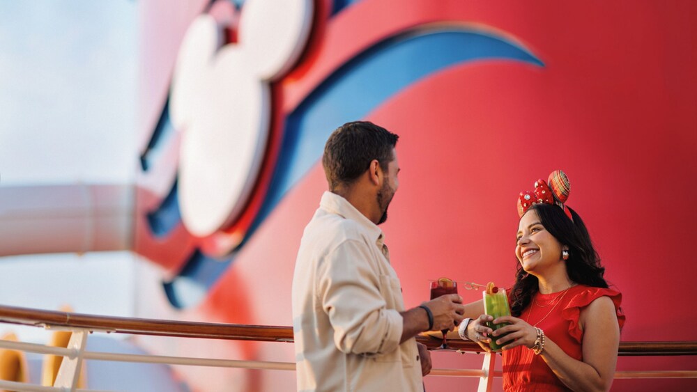 A man and woman holding cocktails while leaning on an upper deck railing