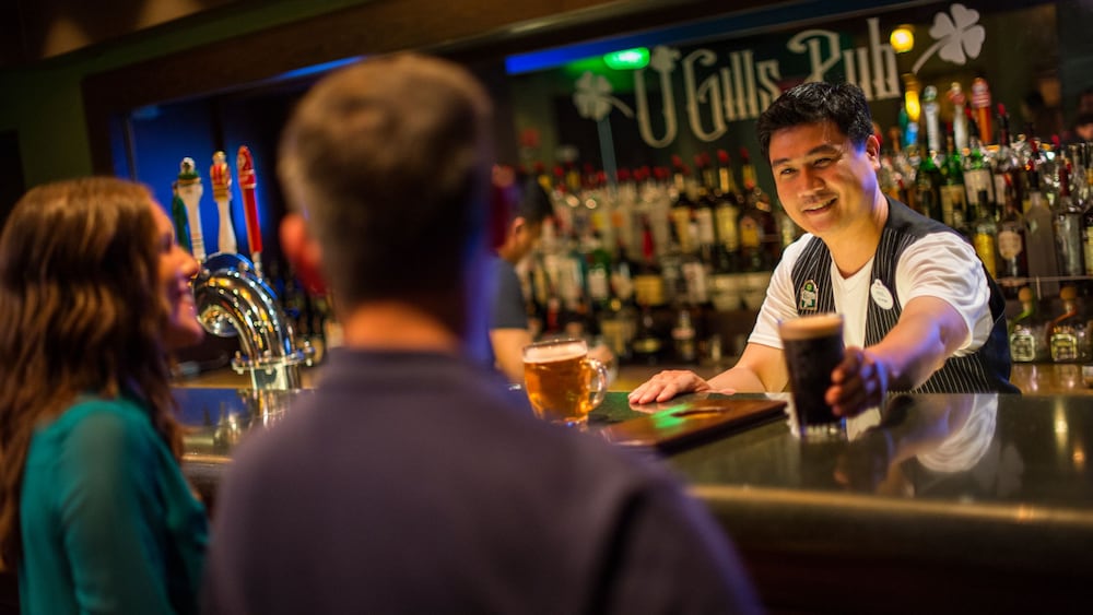 A bartender in a vest serves a beer to Guests seated at the bar in O'Gills Pub
