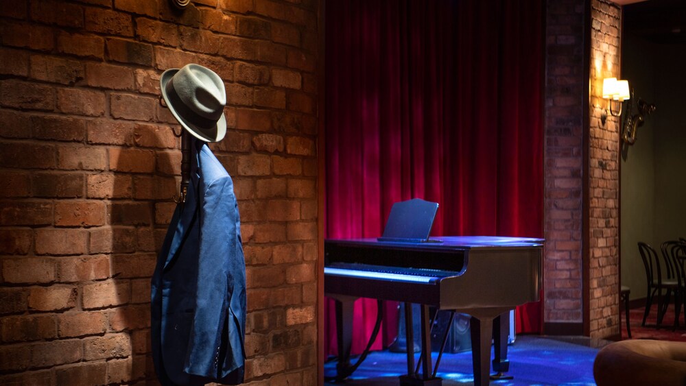 A small stage with a piano and a coatrack with a suit jacket and fedora in the Soul Cat Lounge aboard the Disney Magic