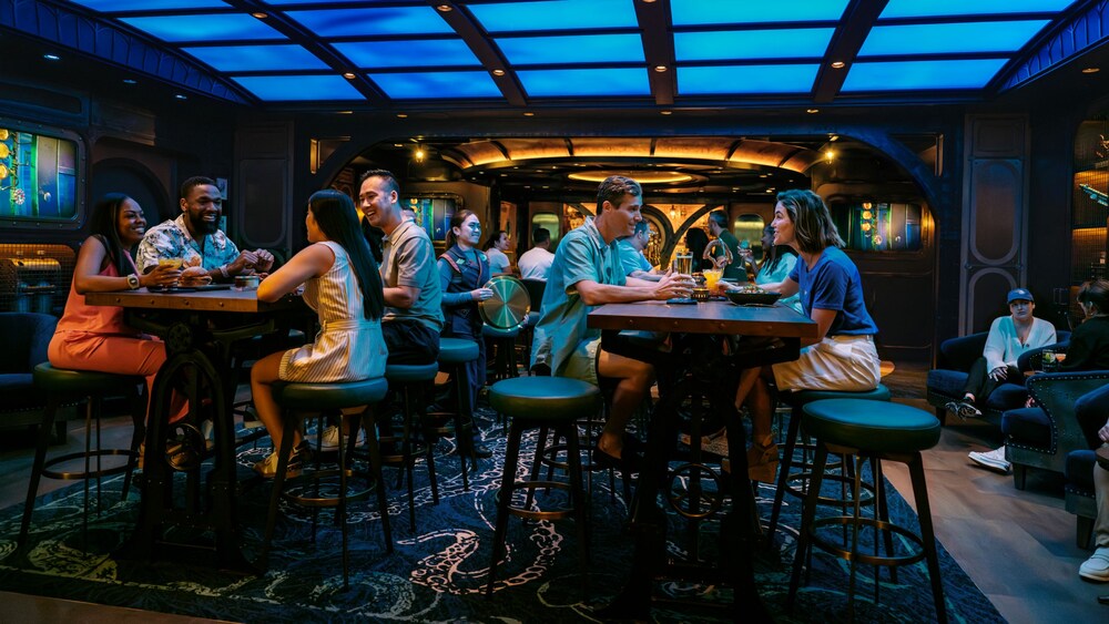 Huéspedes disfrutando de comida y bebidas en las mesas del Periscope Pub, con temática submarina, en el Disney Treasure