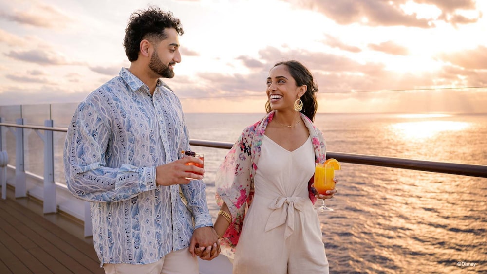 A couple holding hands and drinking beverages while walking on the deck of the Disney Adventure cruise ship